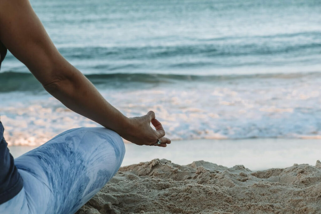 A person mostly offscreen meditating while sitting on sand at the beach.