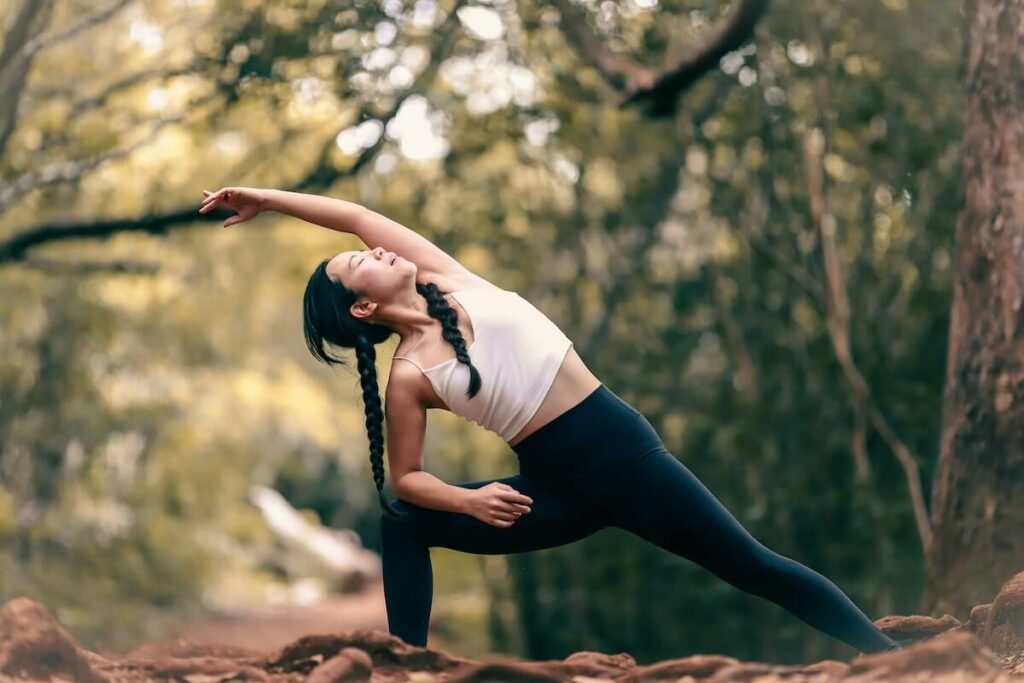 A woman in workout clothes stretching in a wooded area.